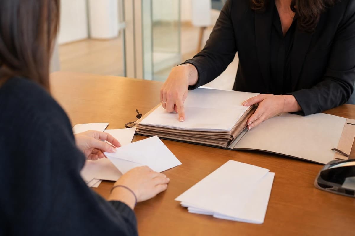 Deux femmes sont assises à une table de bureau et une avocate en tenue sombre montre un dossier papier pendant qu’une…